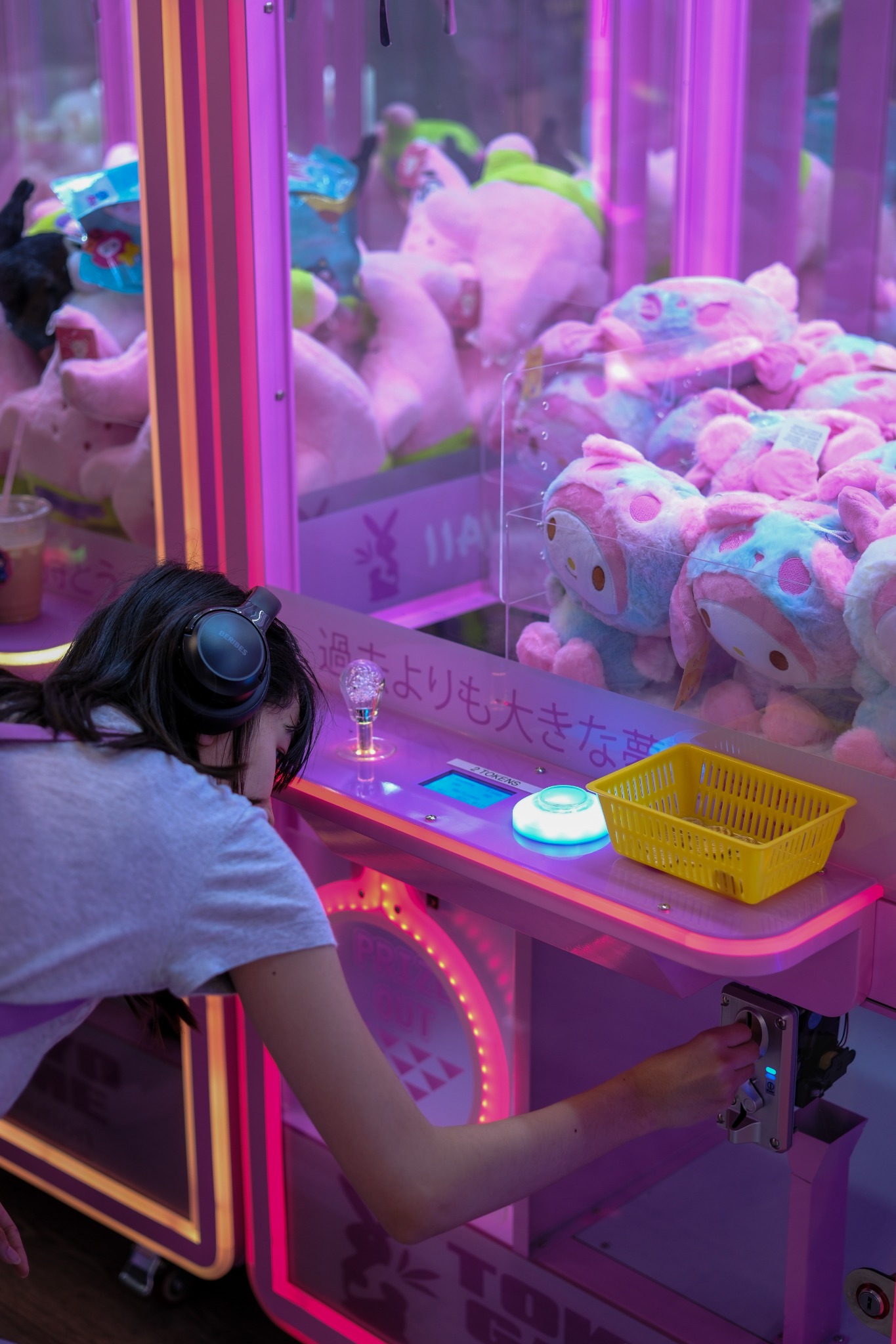 A person wearing headphones inserts a token into a pink claw machine filled with plush toys