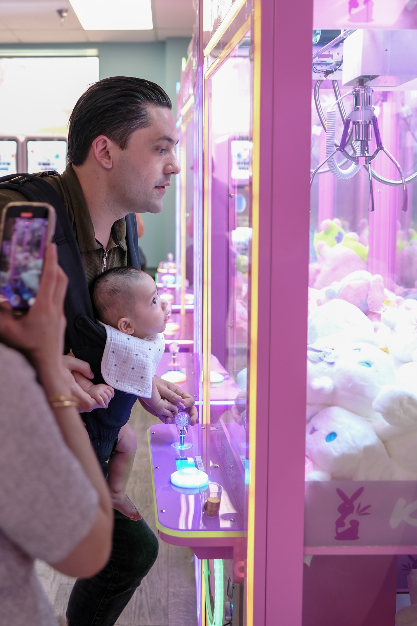 An adult carrying a baby in a front carrier watches a brightly lit pink claw machine