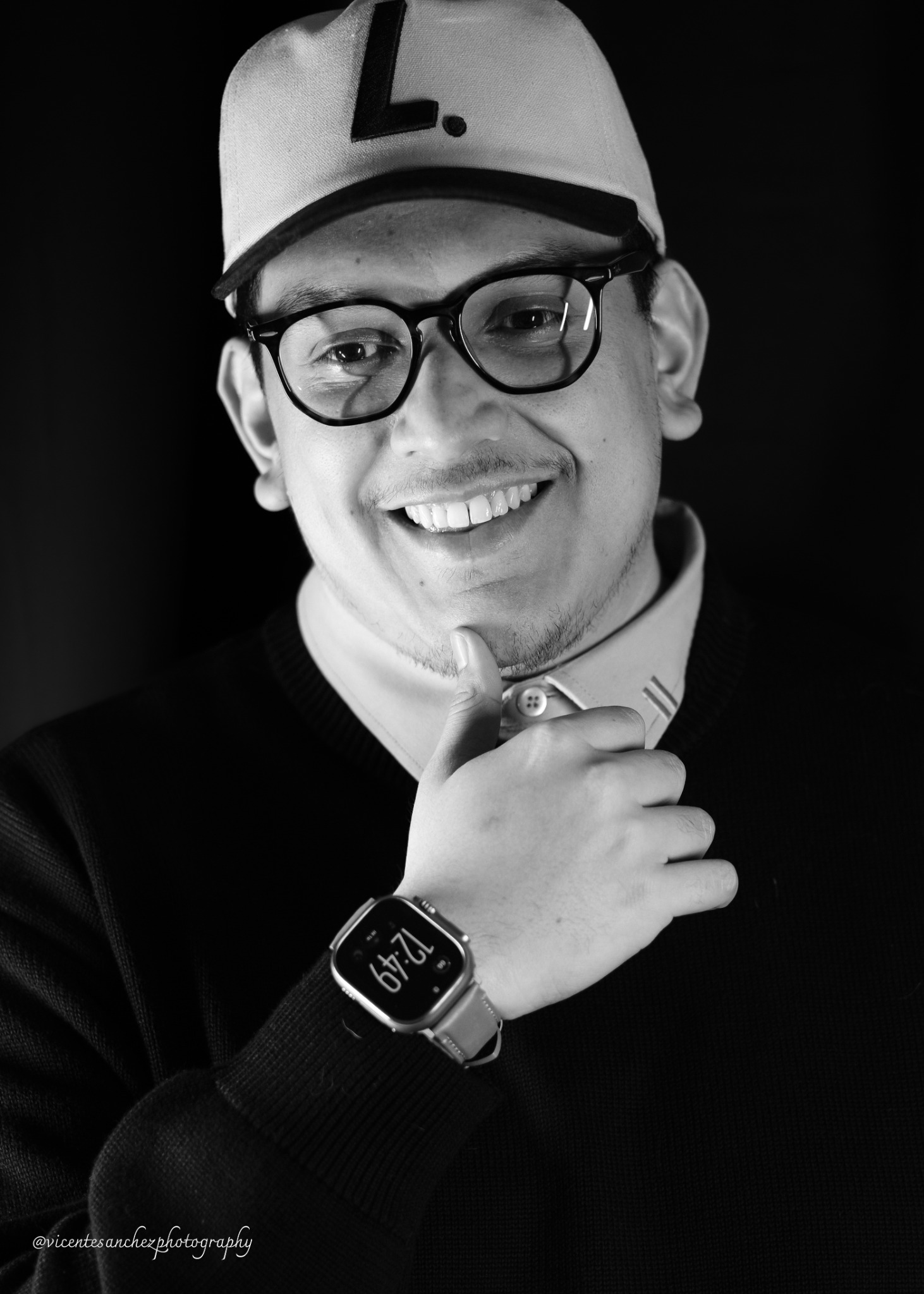 Black-and-white portrait of a smiling man wearing glasses and a baseball cap, with one hand beneath his chin