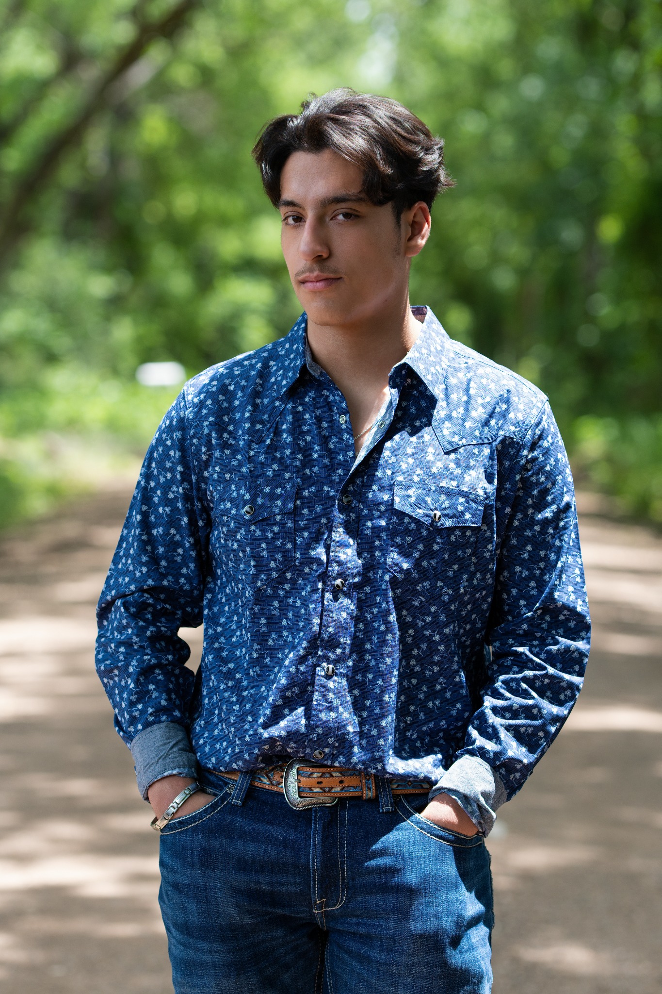 A young man in a blue patterned shirt standing with his hands in his pockets along a tree-lined path