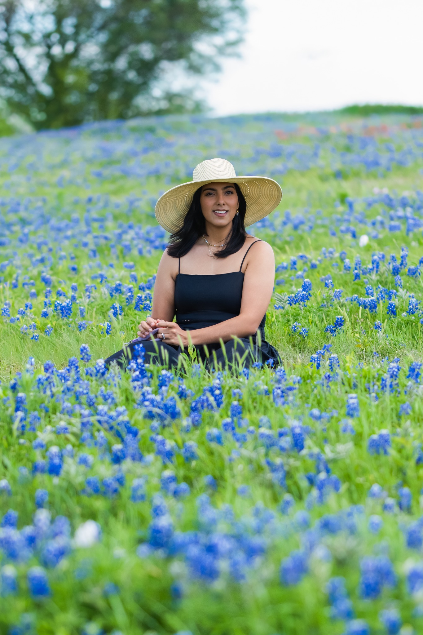 A smiling woman in a sun hat seated among bluebonnets in a green field