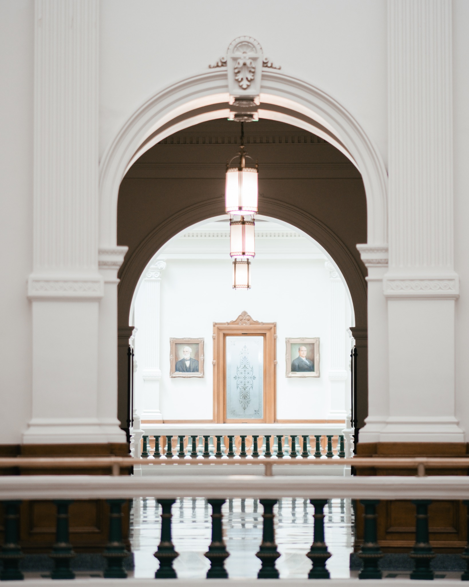 A view through an ornate archway toward hanging lights, a portrait, and a balustrade