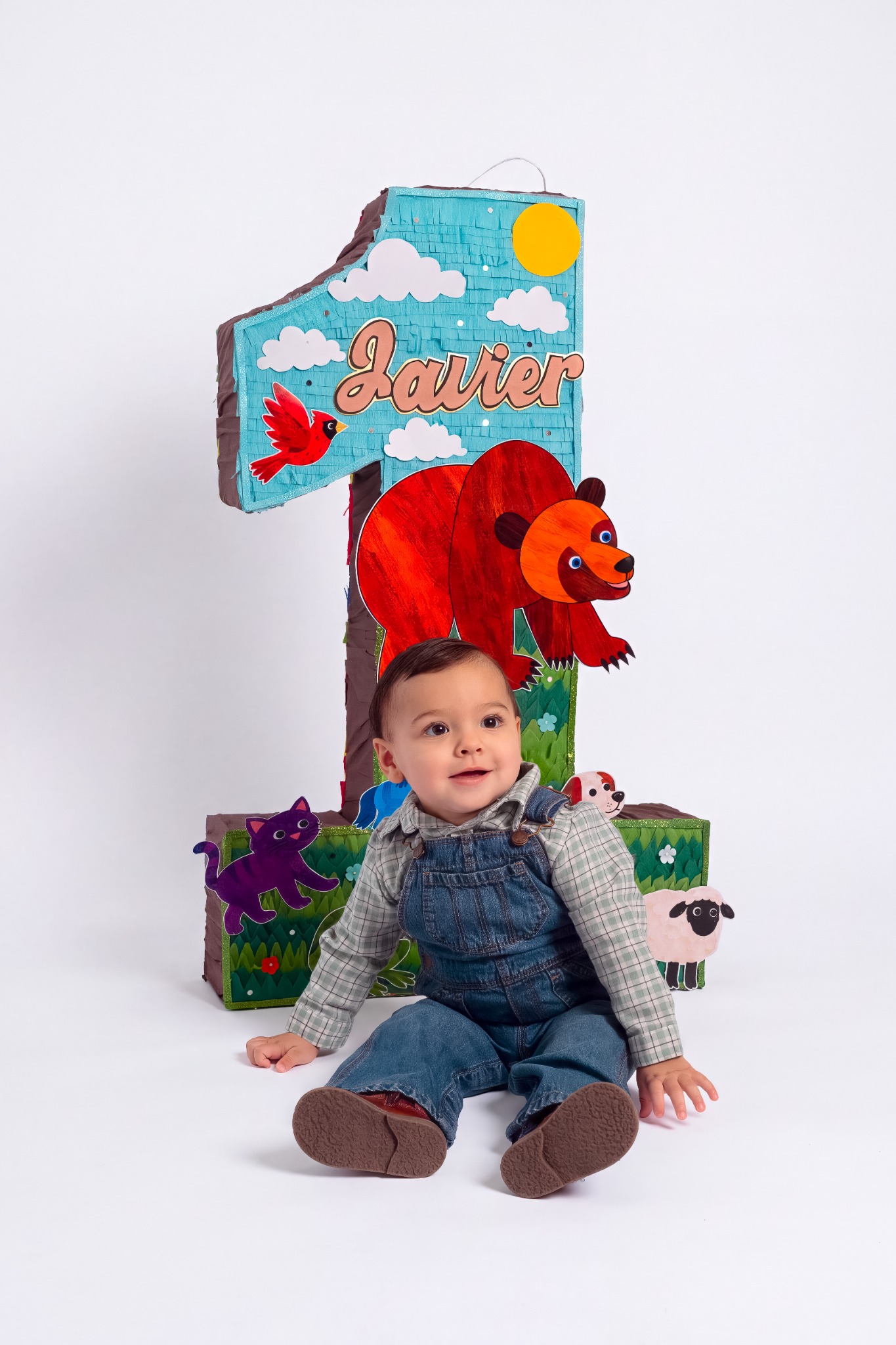 A smiling child in denim overalls seated in front of a colorful number one birthday decoration