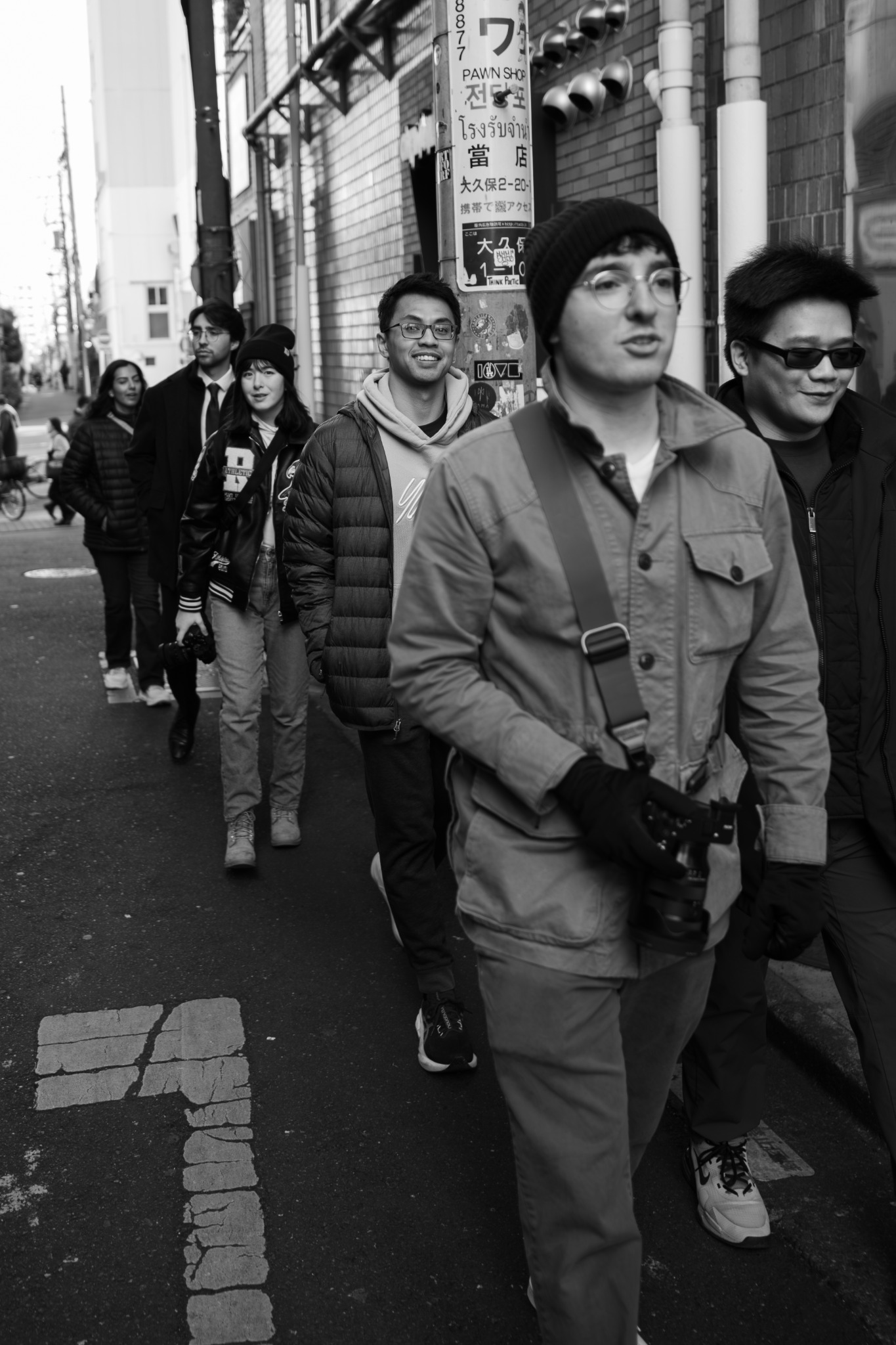 A group walking along a city sidewalk, with a camera-carrying person in front, in black and white