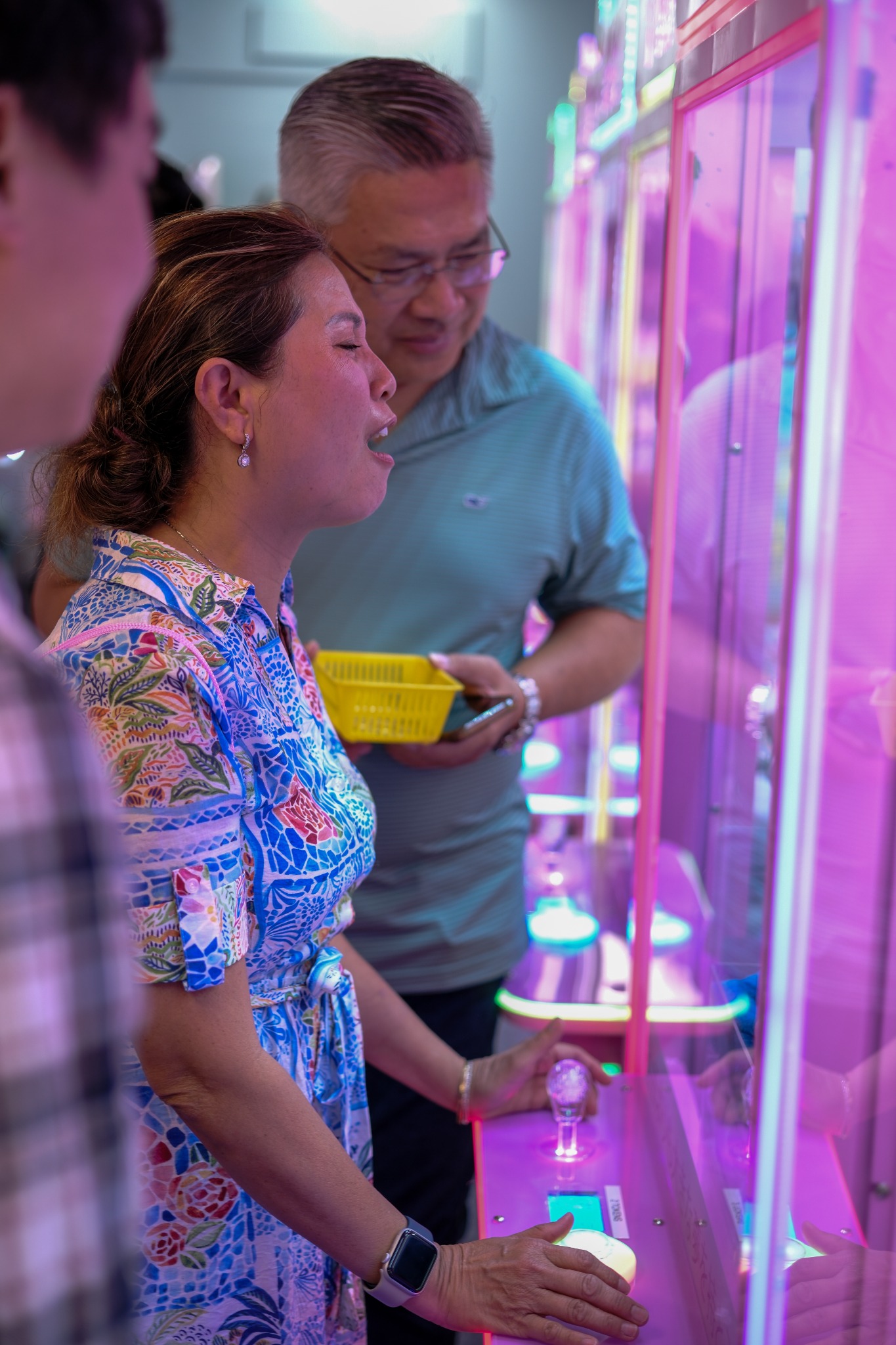 A woman reacts while playing a pink claw machine as a man watches beside her