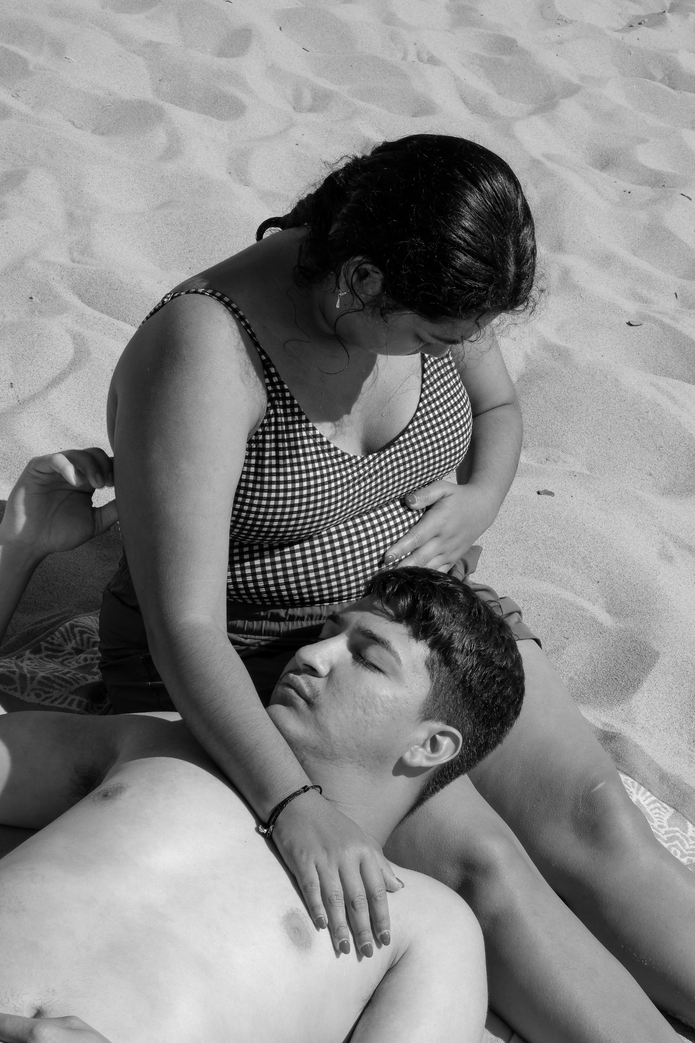 A couple resting together on a sandy beach, with one person's head in the other's lap, photographed in black and white