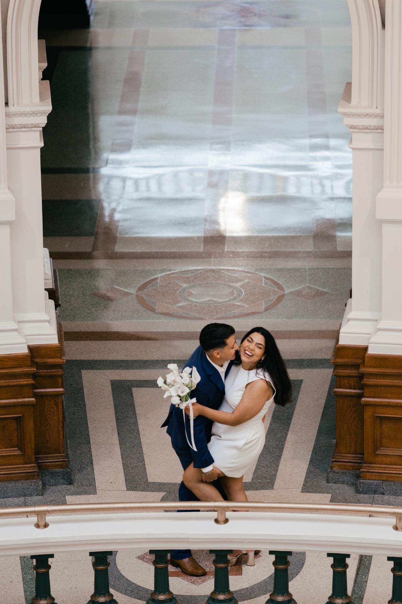 A couple embracing beside an ornate staircase, with the woman in a white dress holding a bouquet