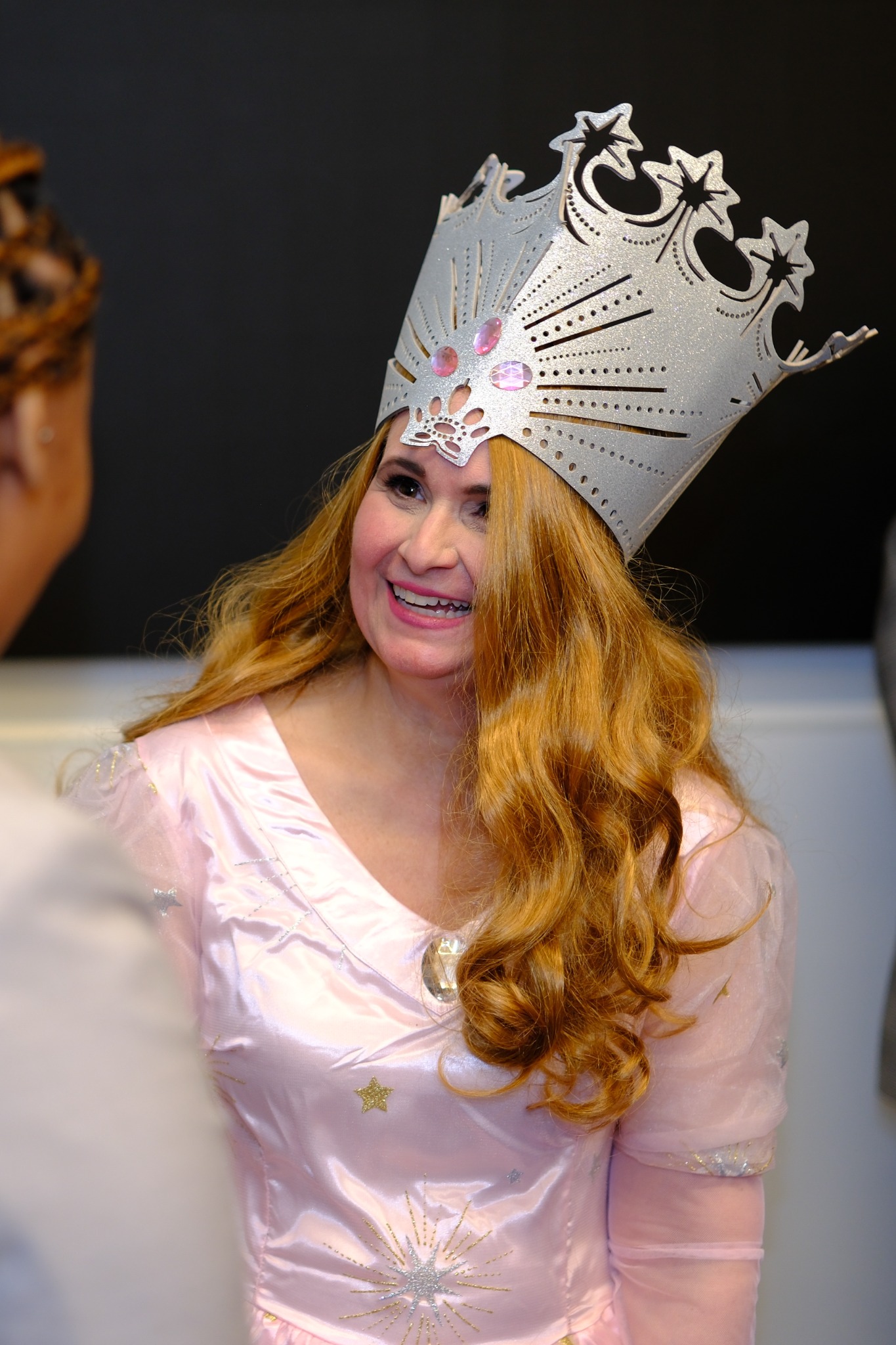 A smiling woman in a pink costume and tall silver crown talking with another guest
