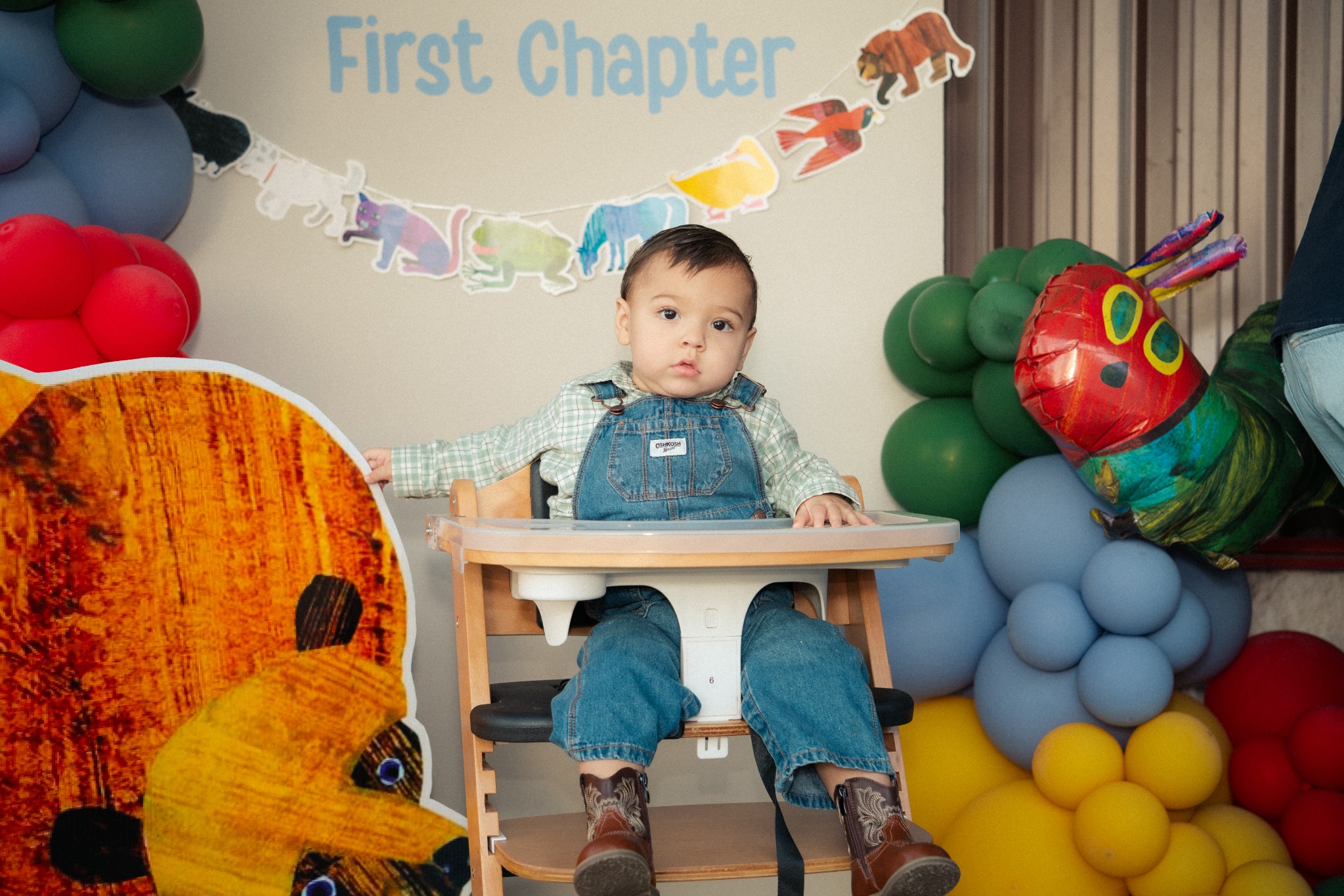 A child in denim overalls seated in a high chair among colorful balloons and birthday decorations