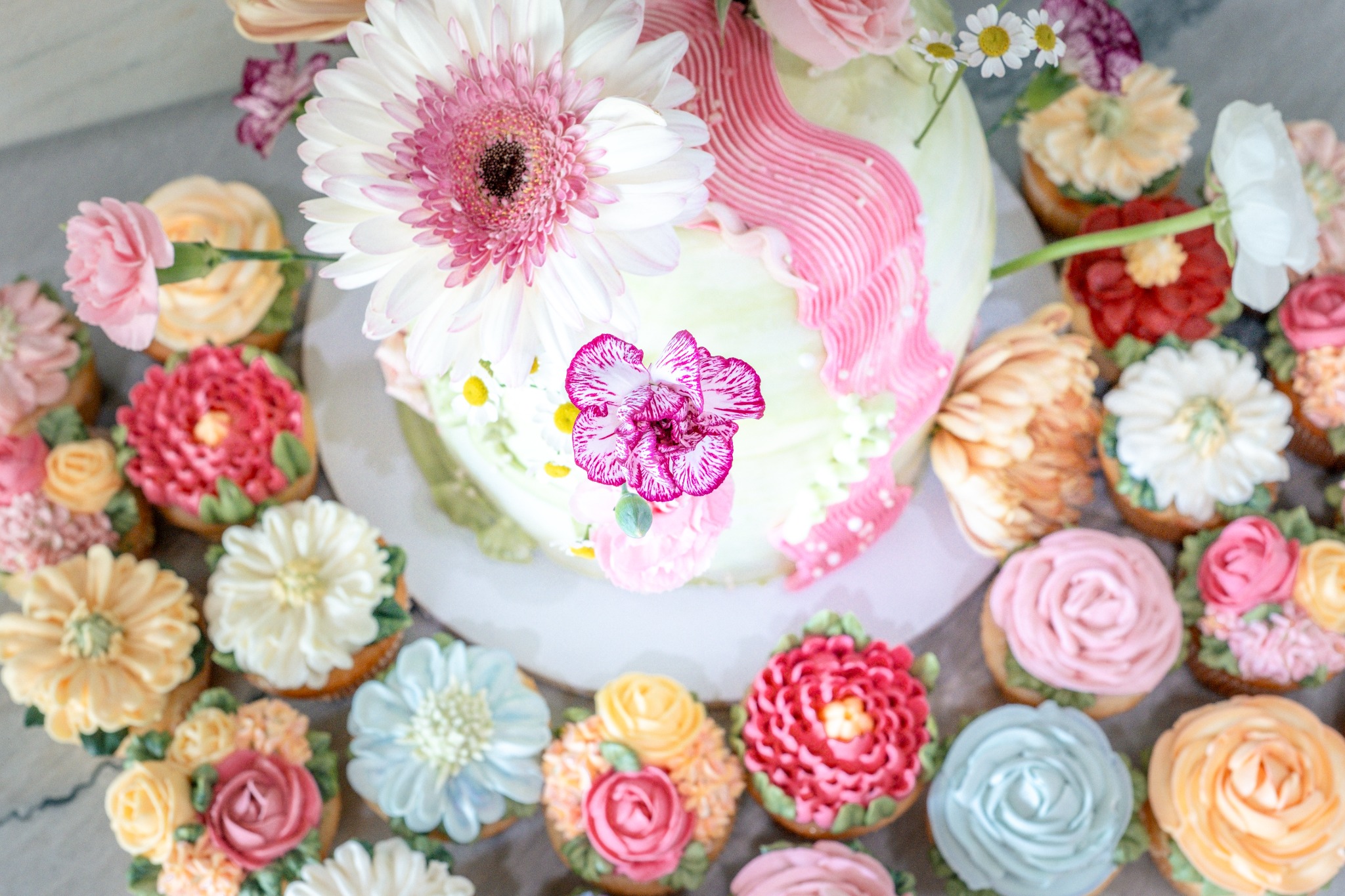 An overhead view of a pastel cake decorated with fresh flowers and surrounded by flower-shaped cupcakes