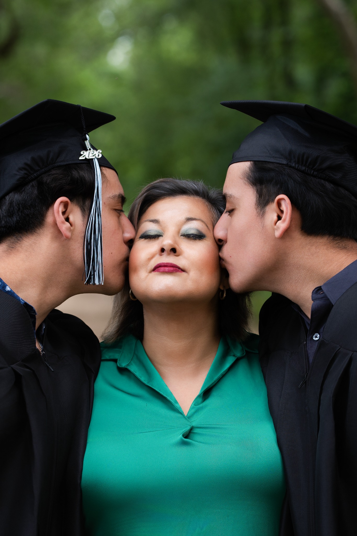 Two graduates in black caps kissing a woman in a green blouse on her cheeks