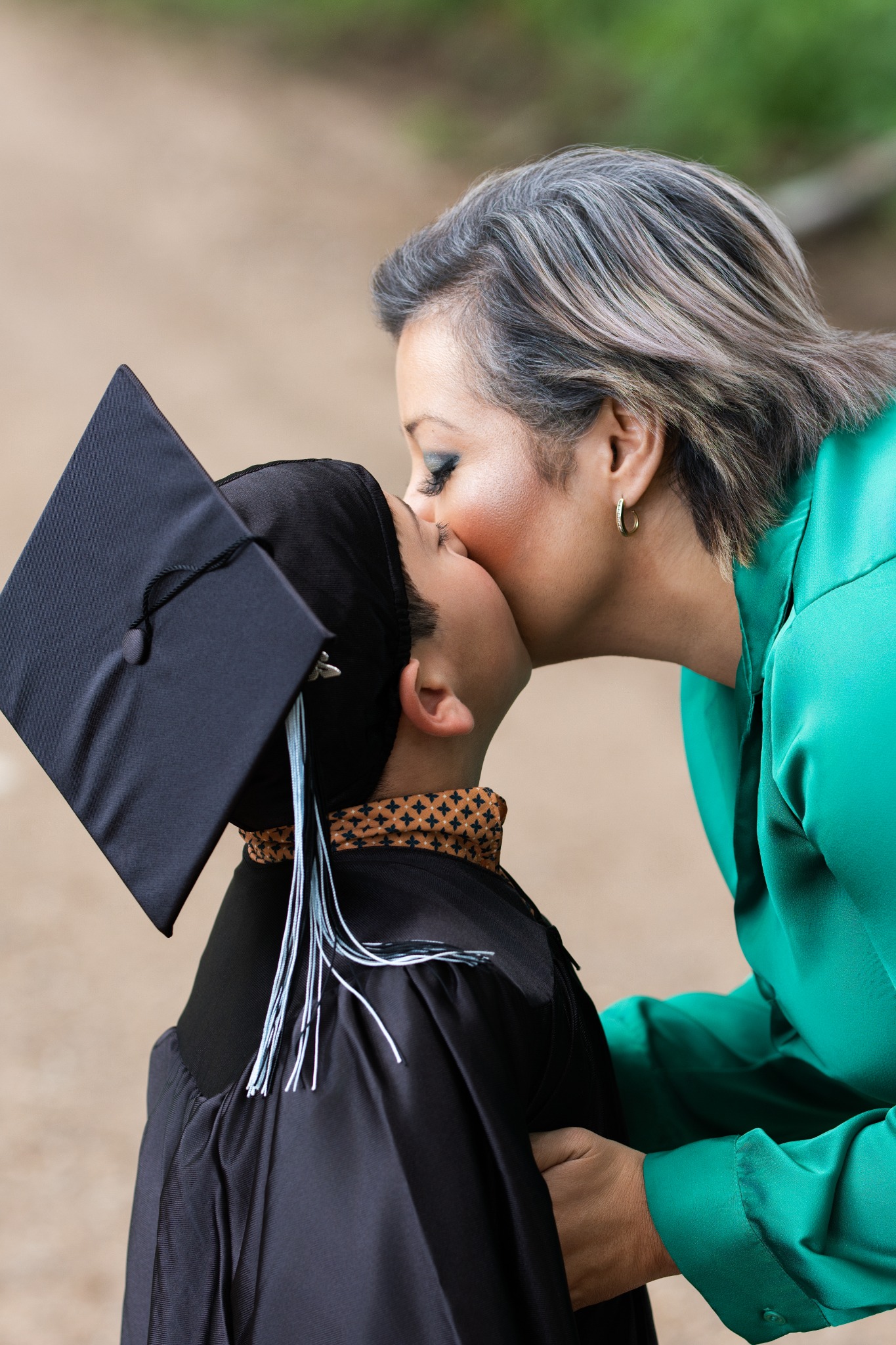 A woman in a green blouse leaning down to kiss a graduate wearing a black cap and gown