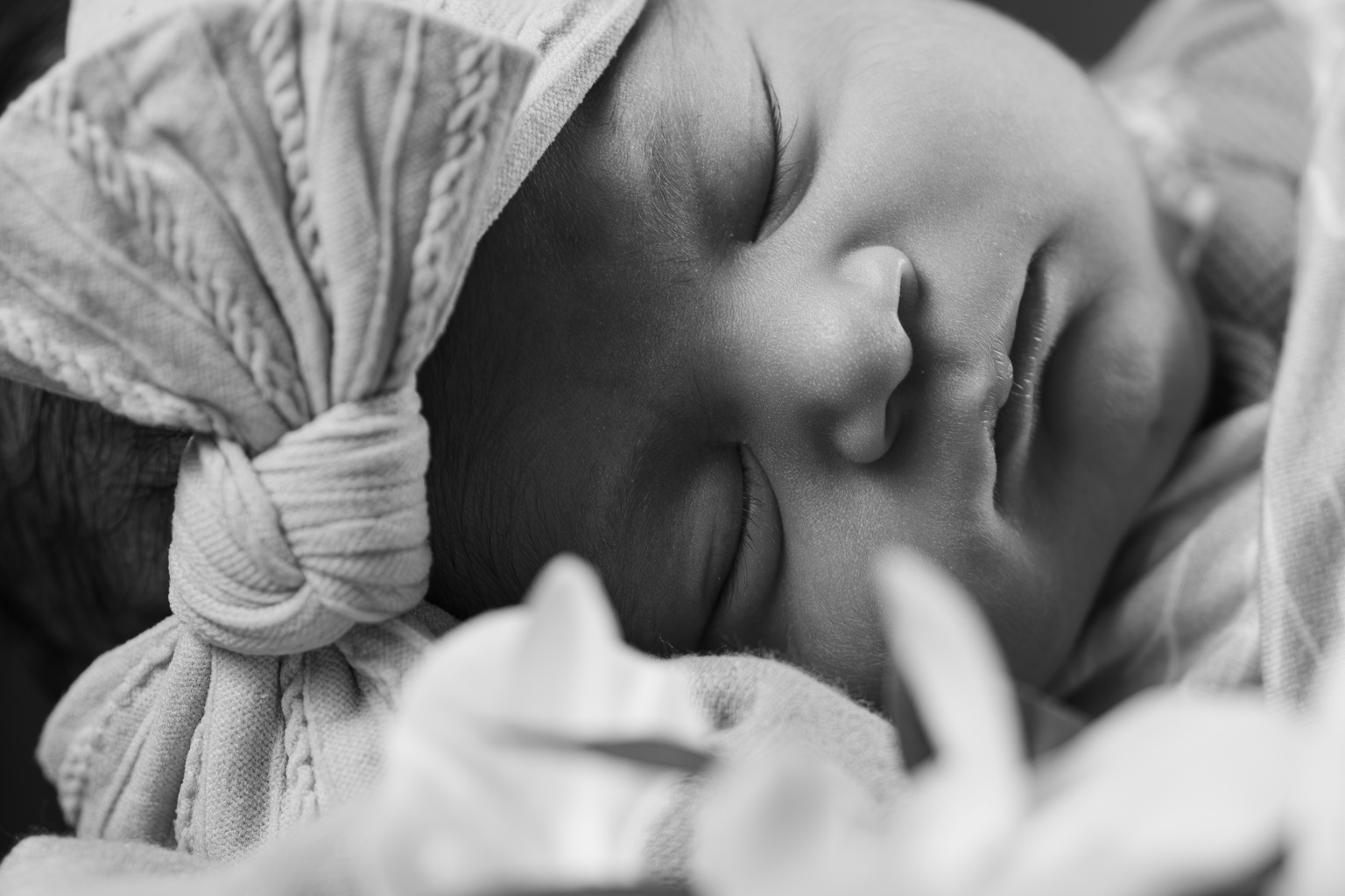 Black-and-white close-up of a sleeping newborn wearing a bow headband, with soft flowers in the foreground