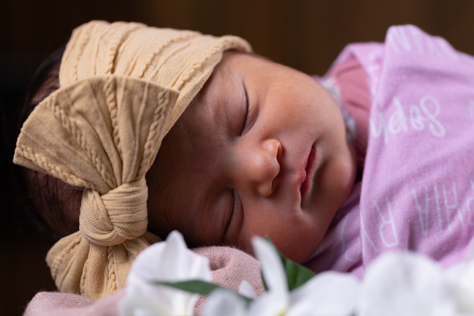 A sleeping newborn wearing a cream bow and pink wrap, with softly blurred white flowers in the foreground