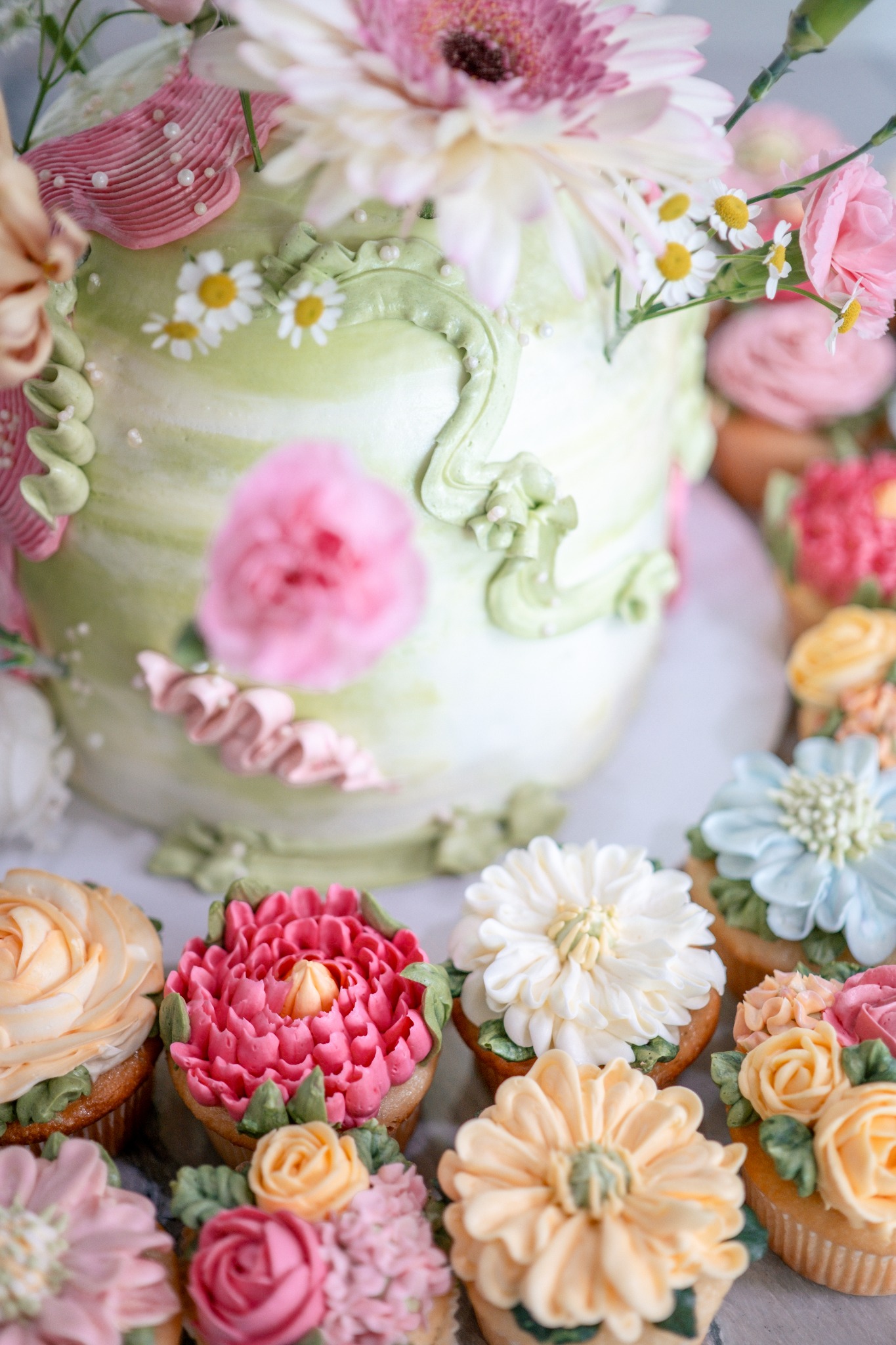 Close-up of a green and pink cake with piped decorations and fresh flowers, surrounded by floral cupcakes