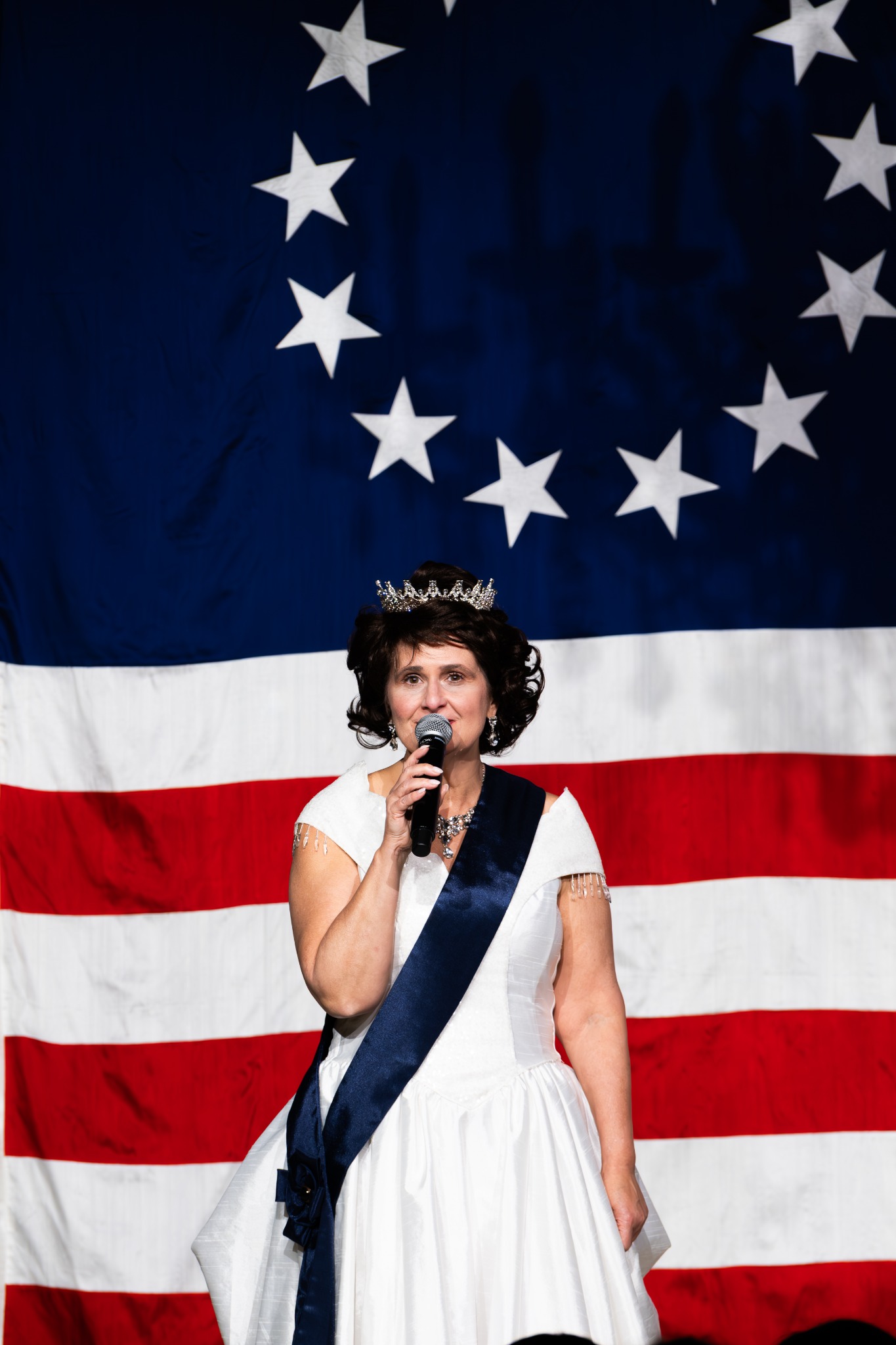 A performer in a white dress, dark sash, and small crown singing into a microphone in front of an American flag