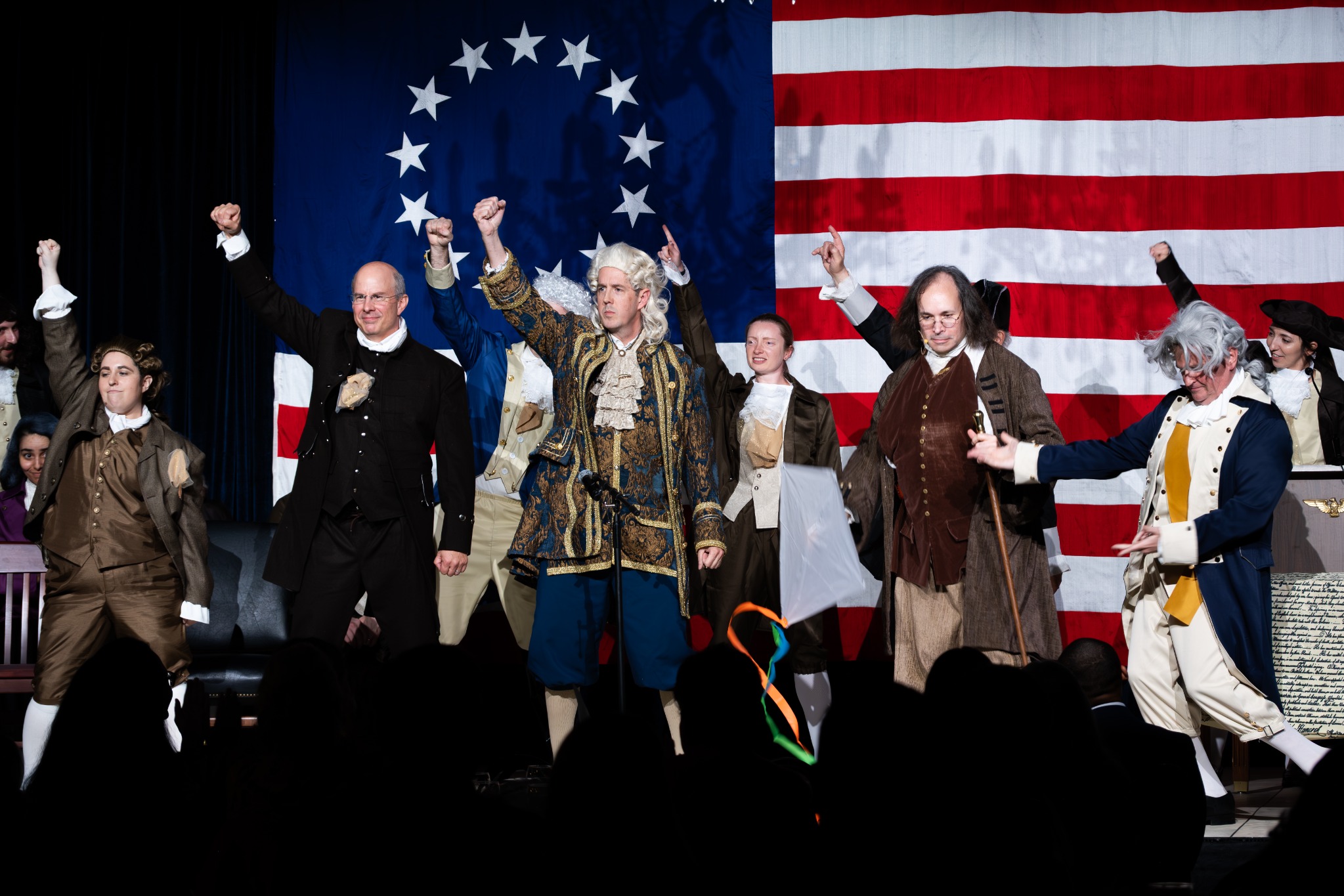 A cast in period costumes performing together in front of a large American flag