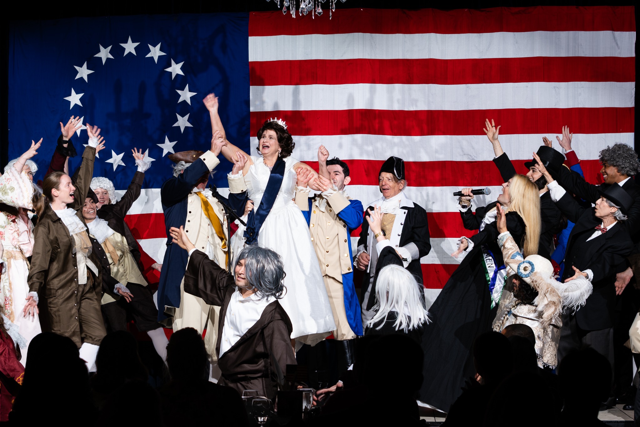 An ensemble in period costumes raising their arms together on a stage backed by an American flag