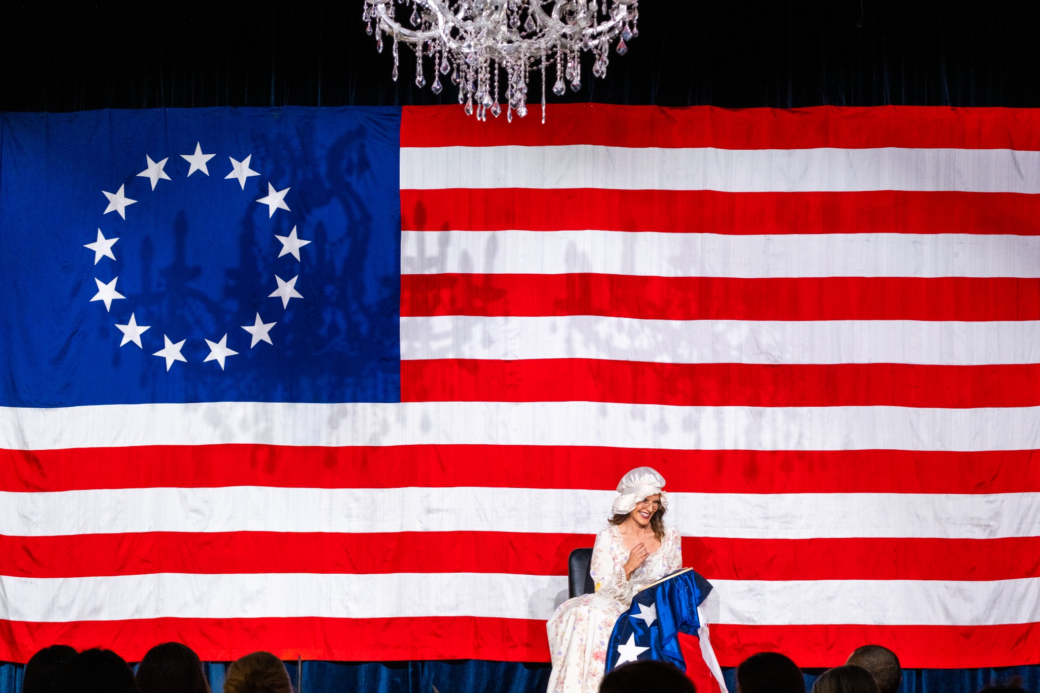 A performer beneath a chandelier, framed by a large American flag