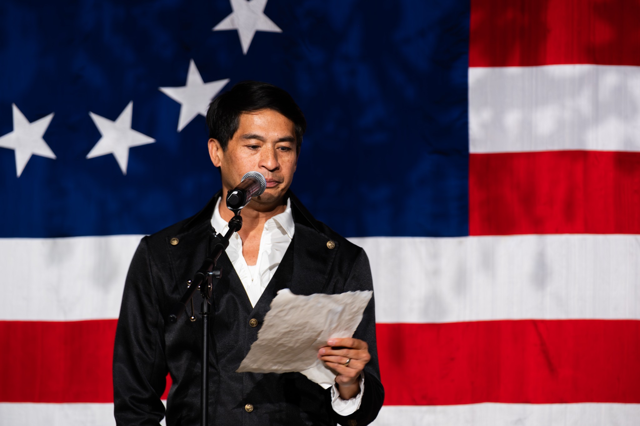 A performer in a dark period jacket reading from a sheet of paper at a microphone