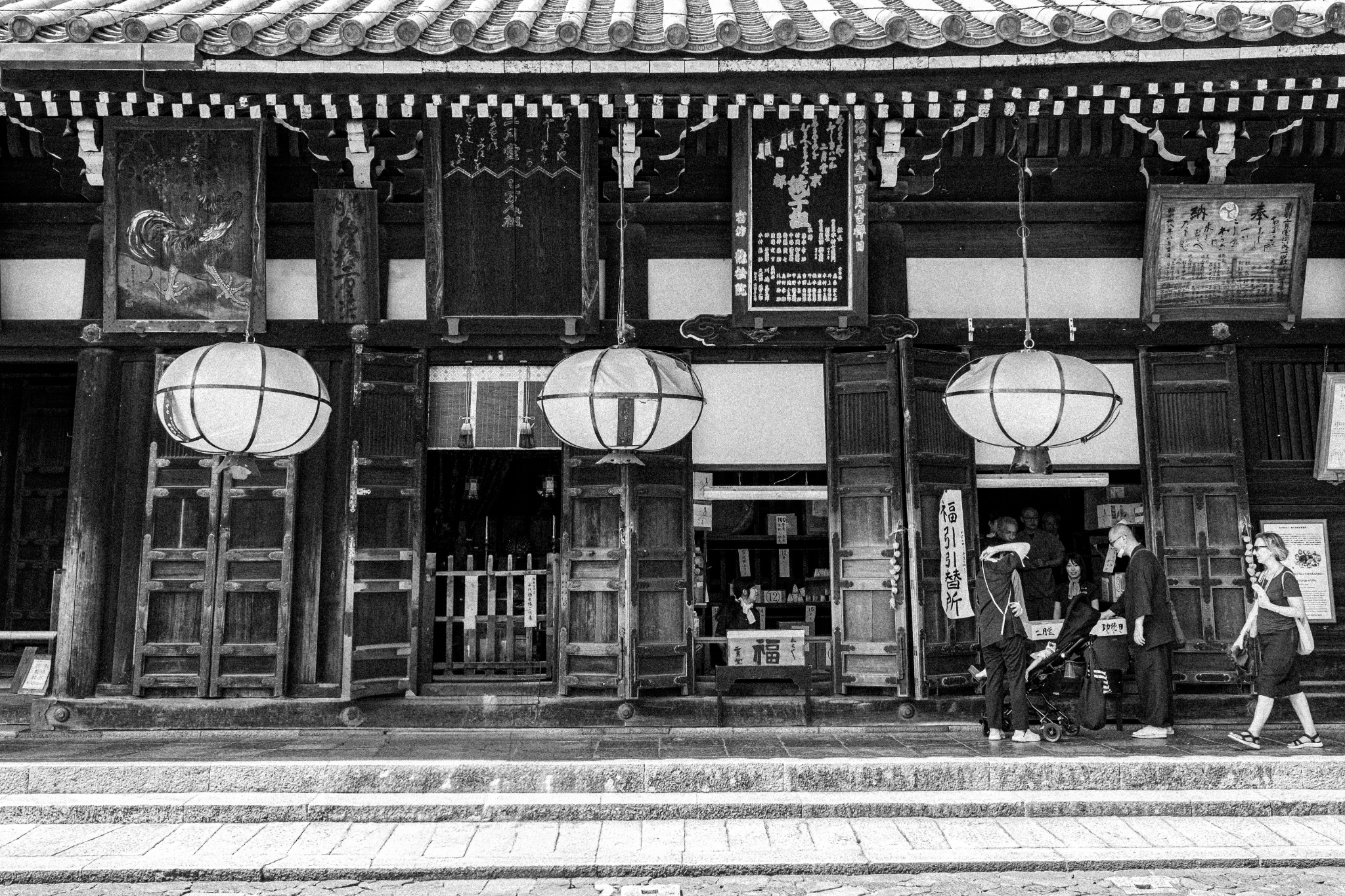 Visitors walking beneath three hanging lanterns along a wooden temple facade, photographed in black and white