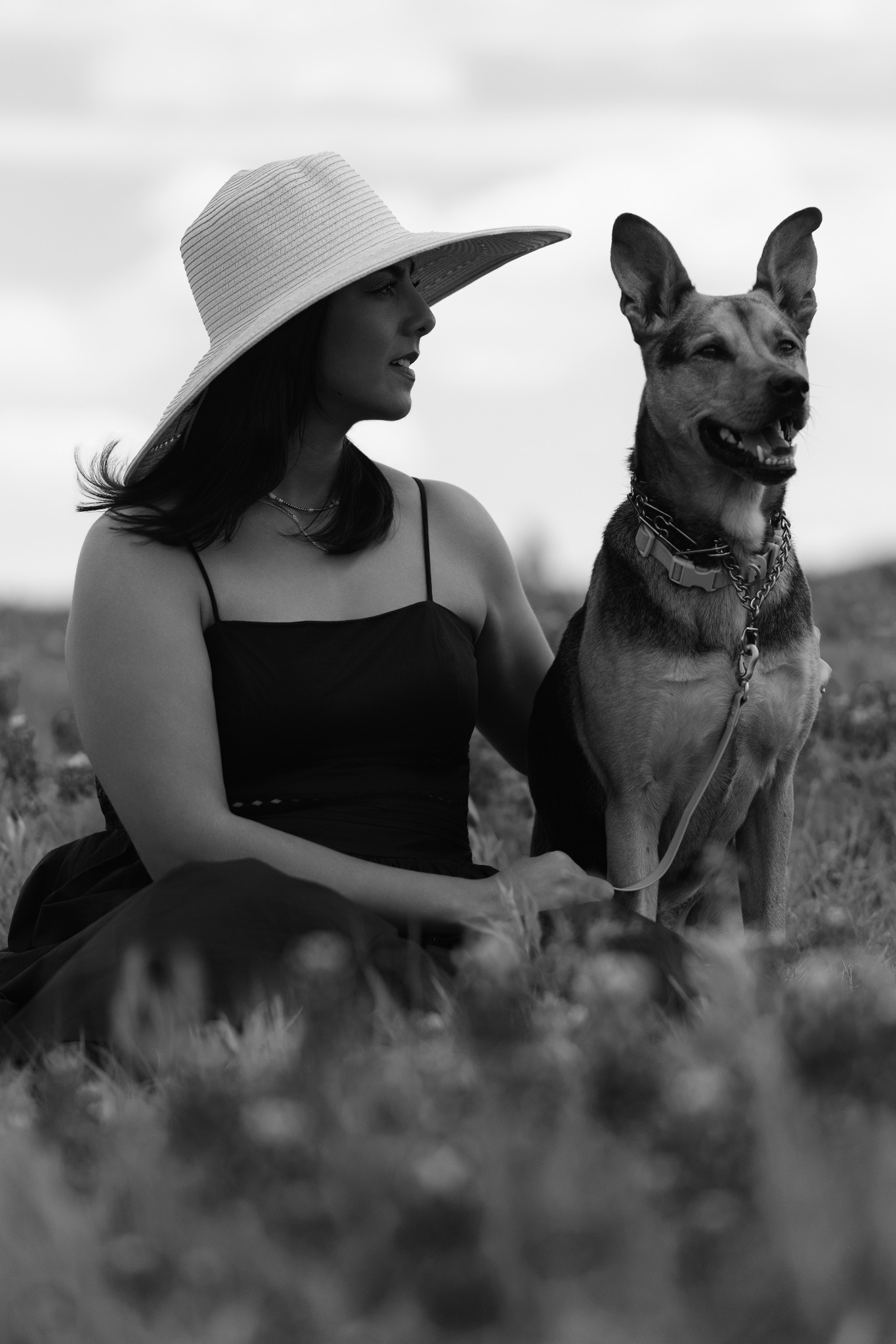 Black-and-white portrait of a woman in a wide-brimmed hat sitting beside a German shepherd